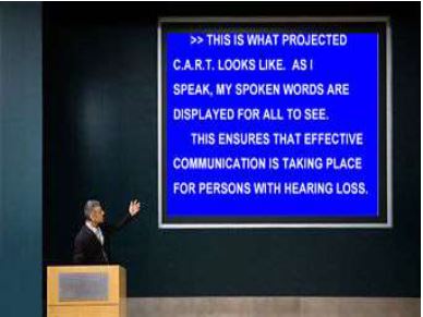 An image displays CART provided for an individual with hearing loss on a large screen at a conference.
